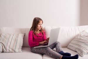 Little girl sitting on sofa with a laptop computer at home. Happy child playing indoors using PC.